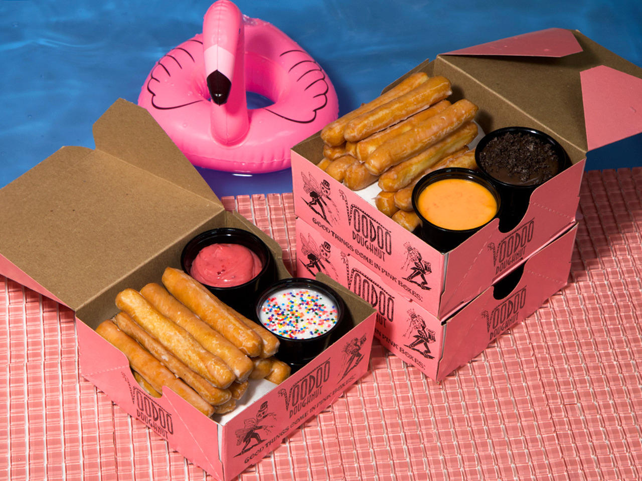 pink tile next to a blue pool with a pink flamingo float inside. On the tile, there are three mini Voodoo Doughnut pink boxes. Two are open and inside are a collection of glazed doughnut fries and dips.